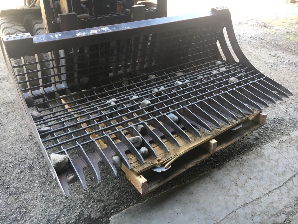 96" skid steer rock bucket with ½" spacing

Heavy-duty skeleton rock bucket on gravel

Close-up of angled bar structure

72" deluxe rock grapple for small rocks and debris

Skid steer lifting rock bucket attachment

